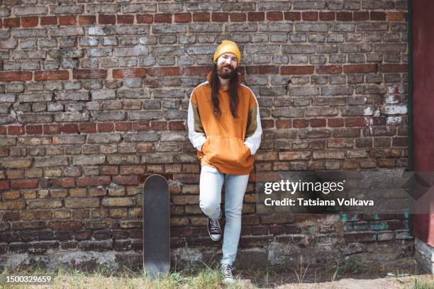 nice bearded male skateboarder with brown hair and beard in orange hoodie standing leaning to brick wall - brown hat 個照片及圖片檔