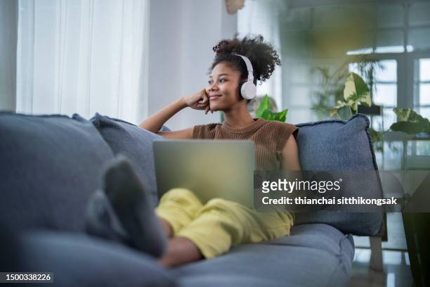 young black woman with wireless headphones listening to music while laying on the sofa at home. - radio stockfoto's en -beelden