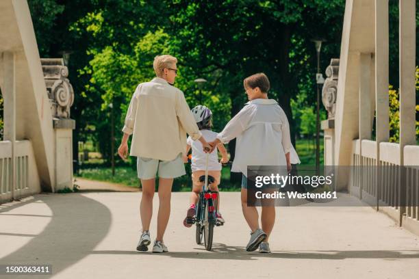 lesbian couple walking with daughter cycling on footpath at park - lesbische stockfoto's en -beelden