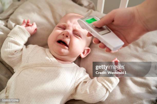mother checking temperature of crying baby lying in crib at home - moms crying in bed stock pictures, royalty-free photos & images