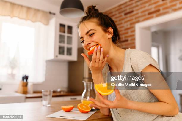 cheerful young woman cutting slices of oranges and eating them for breakfast in the kitchen - woman wearing orange imagens e fotografias de stock