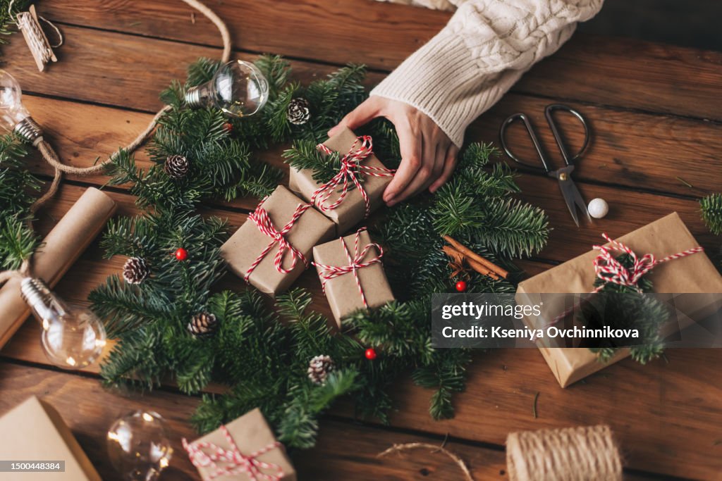 Christmas wreath workshop. Hands holding fir branches, pine cones, berries, thread, scissors on wooden table, flat lay. Making rustic christmas wreath. Authentic rural wreath