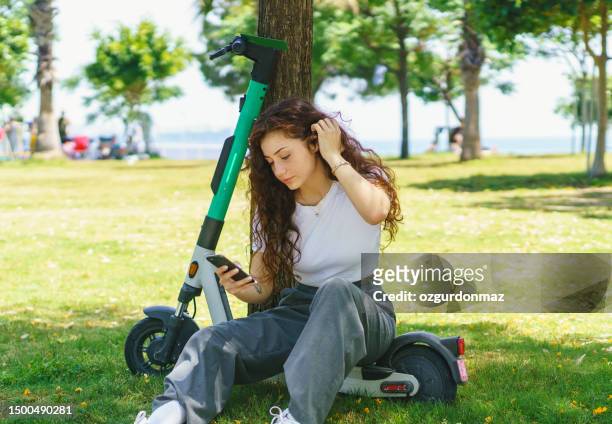 young female parked her electric scooter at park and sitting on the scooter while using smart phone - step well stockfoto's en -beelden