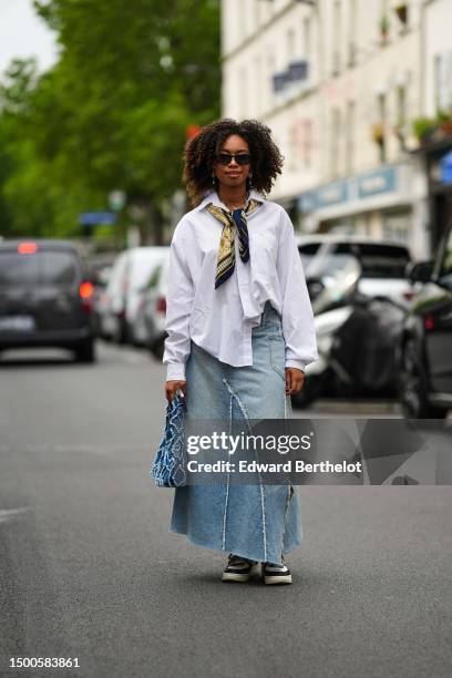 Guest wears black sunglasses, gold snake pendant earrings, a gold large chain with shell pendants necklace, a navy blue with white and gold print...