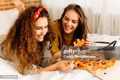 Two girls lying in bed , eating pizza and having fun Two girls lying in bed , eating pizza and having fun