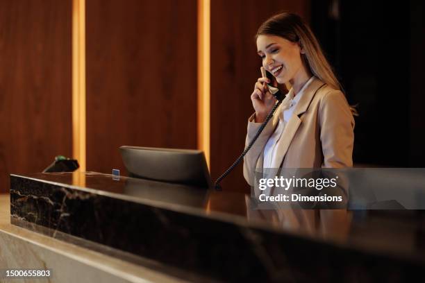 recepcionista caucásica sonriente hablando por teléfono mientras trabaja detrás del servicio de asistencia del hotel - recepcionista hotel fotografías e imágenes de stock