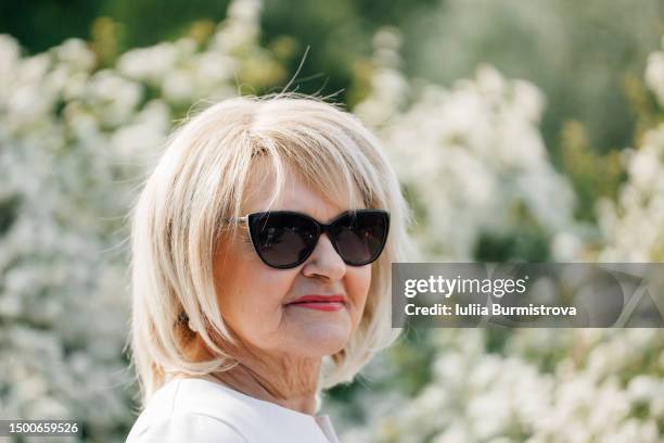 portrait of graceful elderly lady in sunglasses standing in front of blooming bush - frau blond perücke stock-fotos und bilder