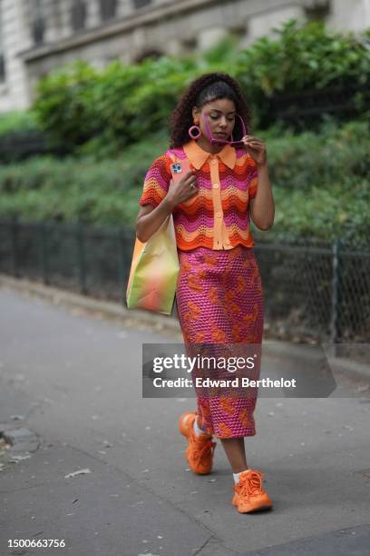 Guest wears neon pink sunglasses, pink earrings, a red / orange / pink wavy striped print pattern braided wool short sleeves / shirt neck buttoned...