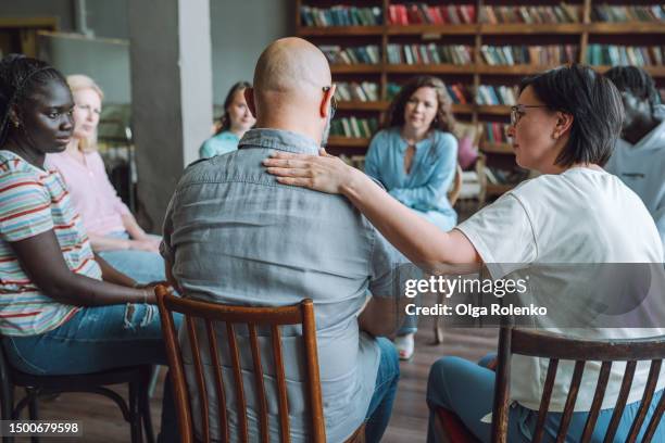 alcohol rehab group session and support. rearview bald man with hand on shoulder sharing problems on psychotherapy session - anonyme alkoholiker stock-fotos und bilder