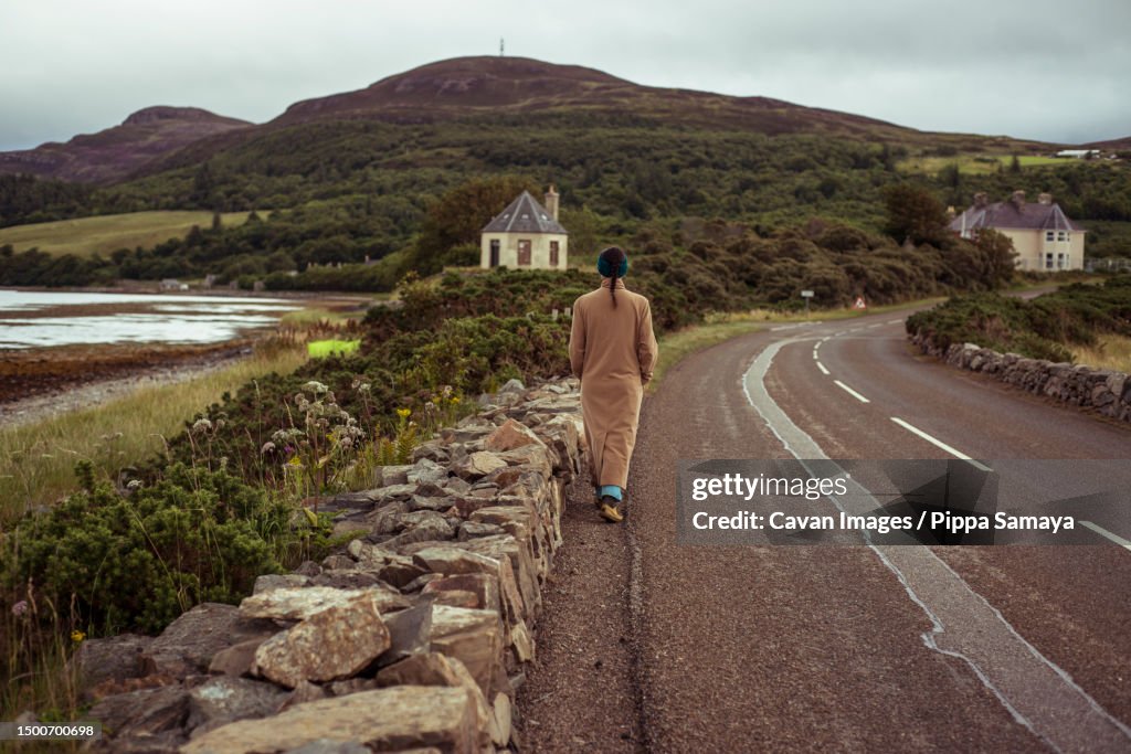 Girl in long jacket walks along remote road in mountains towards town