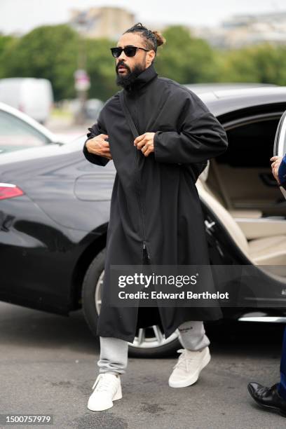 Jerry Lorenzo wears black sunglasses, a black high neck / zipper long coat, pale gray sport pants, white leather laces sneakers , outside Givenchy,...