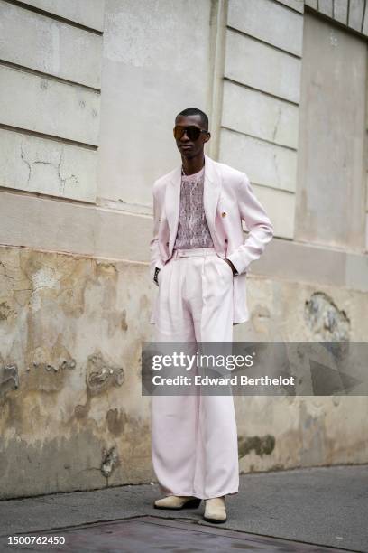 Guest wears black large futurist sunglasses, a pale pink lace print pattern tulle t-shirt, a pale pink blazer jacket, matching pale pink suit pants,...