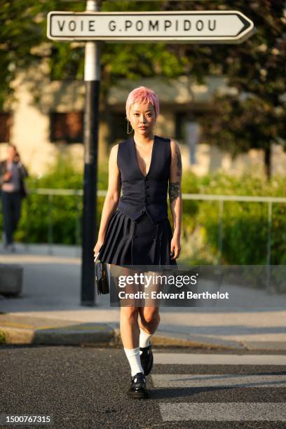 Guest wears gold earrings, a black V-neck / sleeveless / buttoned gilet, a black pleated / accordion short skirt, a black shiny leather with...