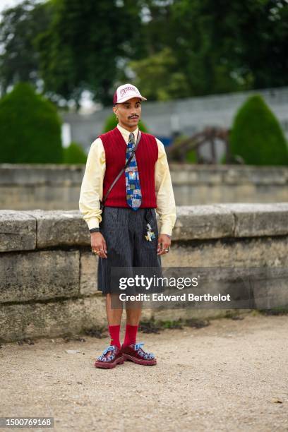 Guest wears a red and white print pattern cap, silver earrings, a pale yellow shirt, a red ribbed wool sleeveless pullover, a royal blue and gray...
