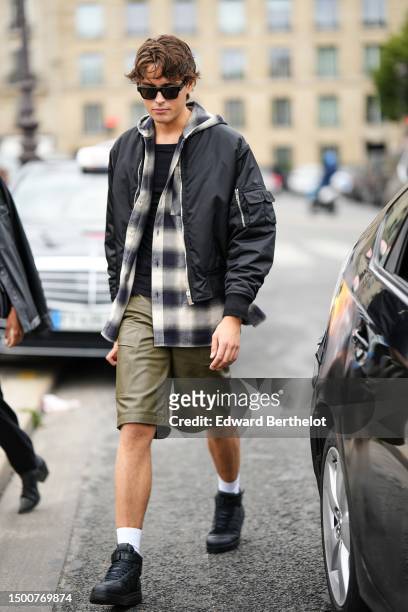 Guest wears black sunglasses, a black t-shirt, a black and white checkered print pattern faded cotton shirt, khaki shiny leather cargo shorts, white...