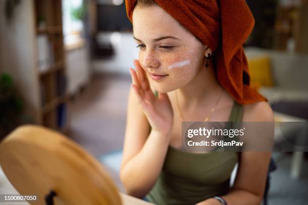 teenage girl, with hair wrapped in a towel, applying face cream in front of the mirror. - face cream stock pictures, royalty-free photos & images