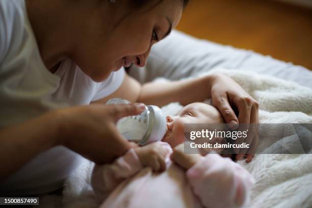 madre sorridente che alimenta la sua neonata con il biberon a casa - nutrire foto e immagini stock