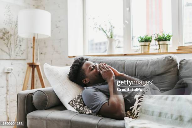 young adult black man lying down at home with a flu and runny nose - blowing nose stock pictures, royalty-free photos & images