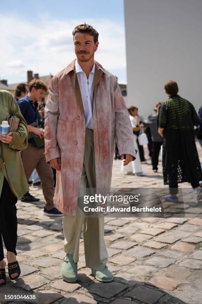 Will Poulter wears a white zipper neck shirt, a pale beige and pink tie and dye print pattern long coat, beige suit pants, pale green matte suede...