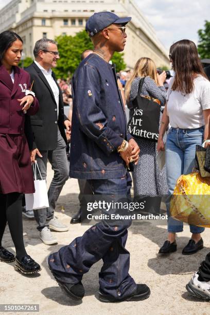 Pharrell Williams wears a navy blue denim with large white embroidered pattern cap, silver and gold rhinestones small circle sunglasses from Louis...