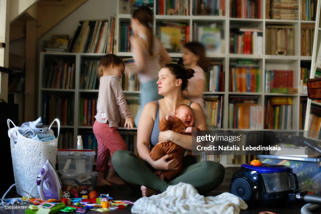 Capturing Maternal Burnout: Overwhelmed Mother Sitting on the Floor in the Living Room amidst Screaming Baby, Sibling Children's Activities, and Household Disorder