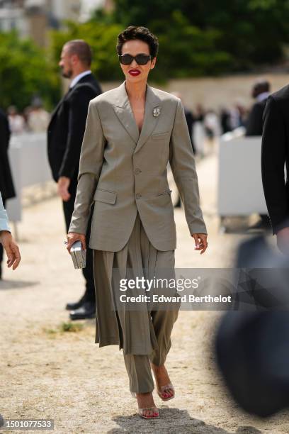 Guest wears black sunglasses, a gray long blazer jacket with a diamonds brooch, pale gray pleated accordion skirt train / suit pants from Dior,...