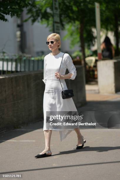 Guest wears black aviator sunglasses, a white long hair midi dress, a black shiny leather shoulder bag from Chanel, black suede pointed slingback...