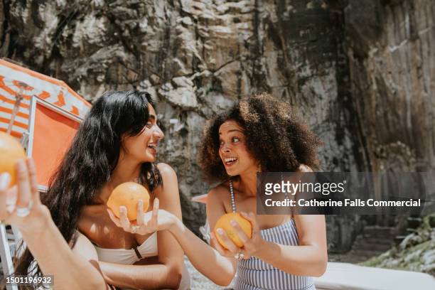 a woman shows her friend oranges, as they sit on a beach - rohkosternährung stock-fotos und bilder