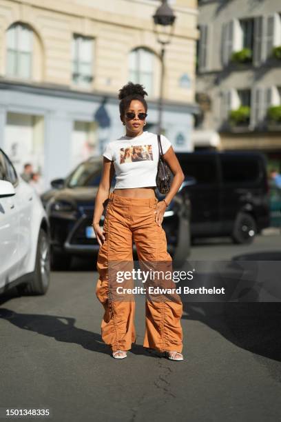 Ellie Delphine wears black circle sunglasses, gold earrings, a gold small chain pendant necklace, a white ribbed with brown print pattern short...