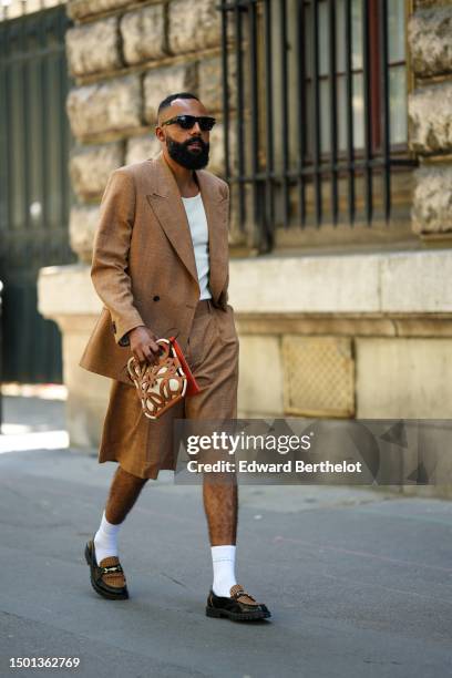 Jean-Claude Mpassy wears black sunglasses, a white tank-top, a brown linen blazer jacket, matching brown suit shorts, white socks, black shiny...