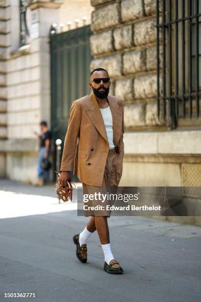 Jean-Claude Mpassy wears black sunglasses, a white tank-top, a brown linen blazer jacket, matching brown suit shorts, white socks, black shiny...