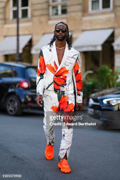Guest wears black squared sunglasses, a gold large chain necklace, a white with neon orange and black large flower pattern blazer jacket, matching...