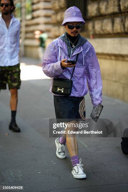 Guest wears a purple nylon bob hat, black sunglasses, a black t-shirt, silver chain necklaces, a gray nylon zipper hoodie rain coat, a purple nylon...