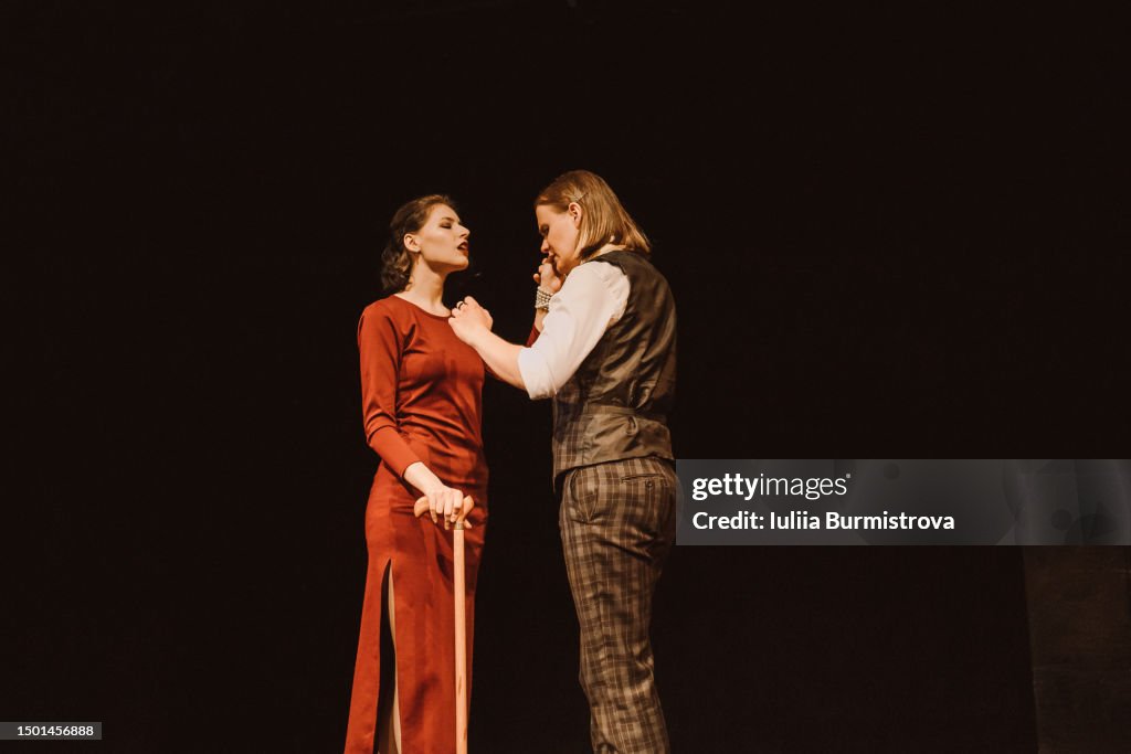 Beautiful woman in red dress and man in vintage suit standing close while performing on stage in college theater club