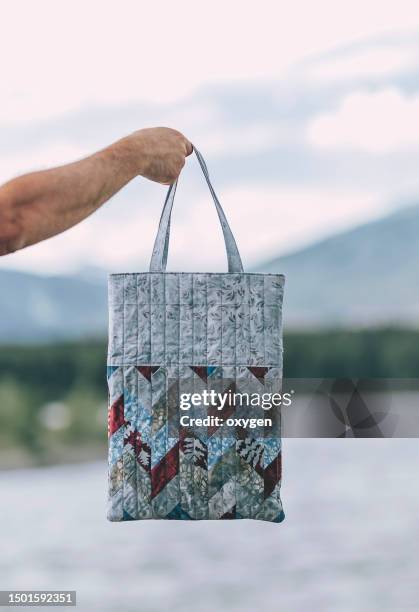 bag held by a hand.  reusable patchwork shopping bag with rhombus shapes. scandinavian hugge style. needlework and quilting. nature mountain and river bachground - トートバッグ 無人 ストックフォトと画像