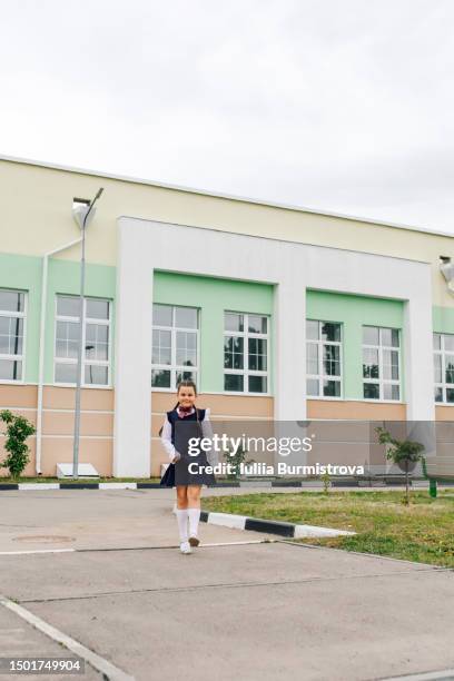 carefree small girl in school uniform walking on sidewalk with school building in background - elementary school building stock pictures, royalty-free photos & images