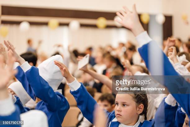 serious girl in blue school uniform sitting in crowd of classmates at school assembly hall with raised hand - assembly room stock pictures, royalty-free photos & images