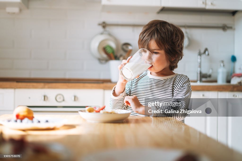 Cute small child boy 4-5 years having breakfast drinking milk in kitchen at home