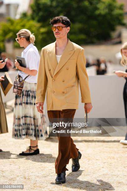 Guest wears brown and orange sunglasses, a white t-shirt, a yellow buttoned long blazer jacket, dark brown velvet suit pants, black shiny leather...