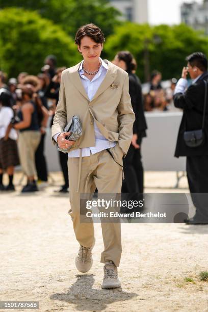 Guest wears a white shirt, a beige blazer jacket from Dior, matching beige suit pants from Dior, a gray monogram print pattern in coated canvas...