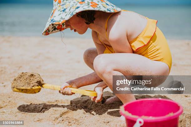 a little girl wearing a wide-brimmed sun hat squats on the beach and digs in the sand - ankle deep in water stock pictures, royalty-free photos & images