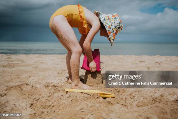 a child, doubled over, uses a pink plastic pail to build a sandcastle - ankle deep in water stock pictures, royalty-free photos & images