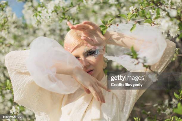 blonde girl in white blouse on blooming cherry tree background. short pink hair. model in skirt. beauty makeup with feather. style and fashion. haute couture - high fashion stock pictures, royalty-free photos & images