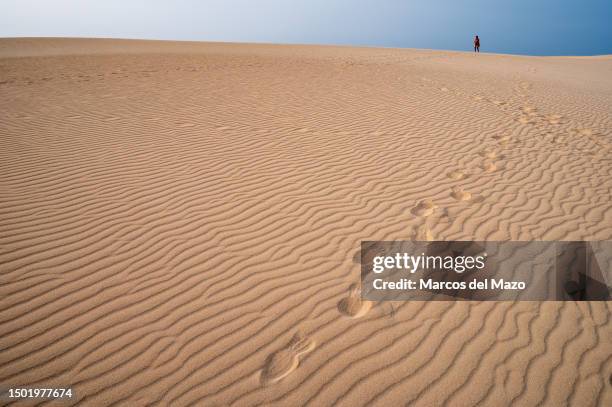 Footprints and a tourist walking along the sand during a hot summer day at the Dunes of Corralejo, north of Fuerteventura in the Canary Islands. The...