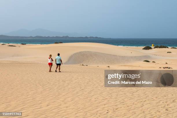 Tourists walk along the sand during a hot summer day at the Dunes of Corralejo, north of Fuerteventura in the Canary Islands. The Corralejo Dunes are...