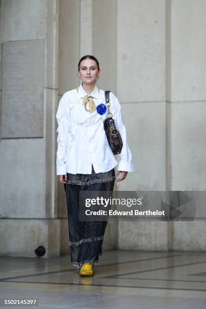 Callisto Van Soeterstede wears a black hair clip, silver earrings, a white pearls small necklace, a white oversized ruffled shirt, a pale yellow with...