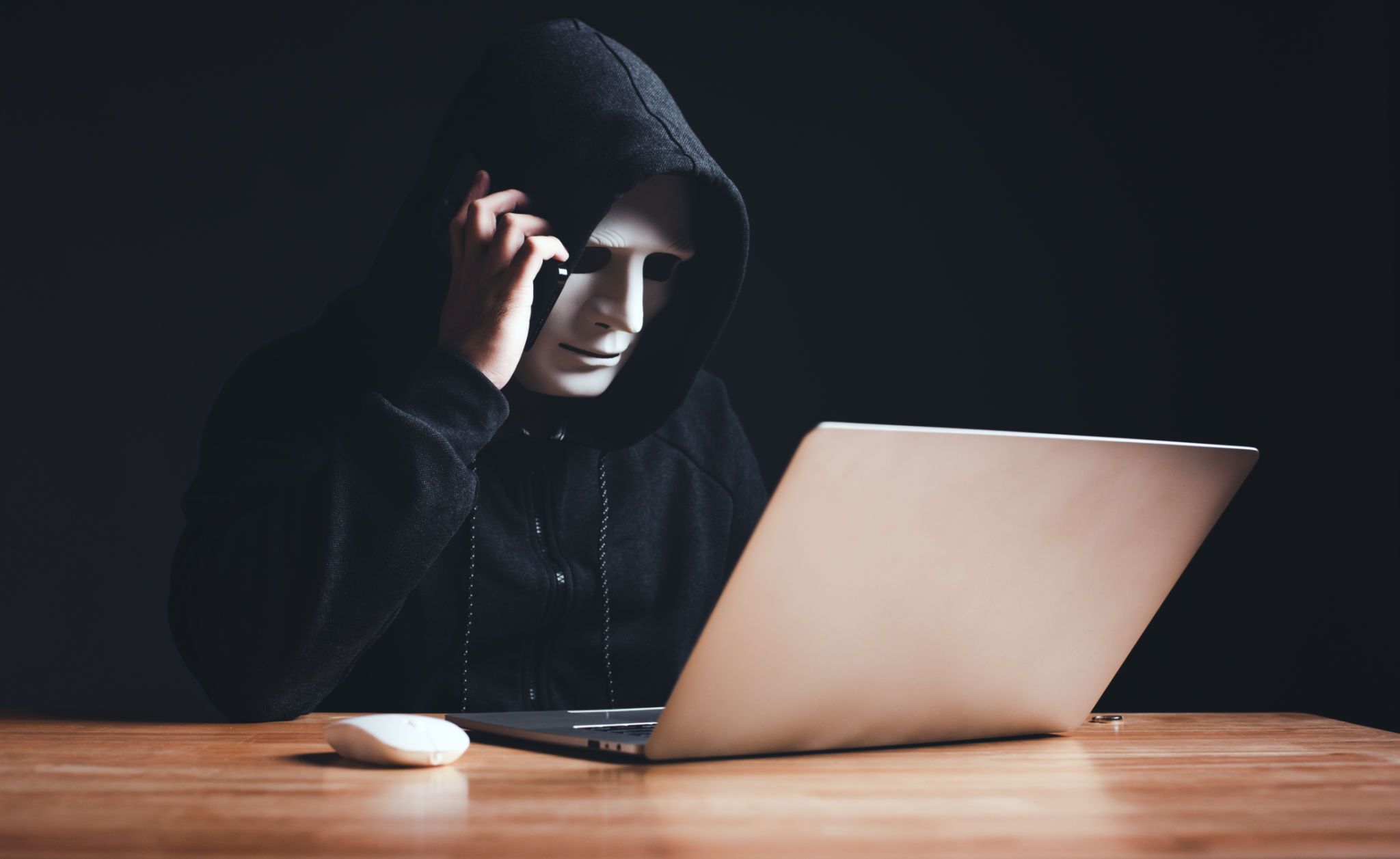 Black hat hacker in hood using laptop computer and call smartphone to victim on desk hacking privacy sensitive data hack in dark room background. Cyber security cyber crime concept. Hacking phishing Black hat hacker in hood using laptop computer and call smartphone to victim on desk hacking privacy sensitive data hack in dark room background. Cyber security cyber crime concept. Hacking phishing