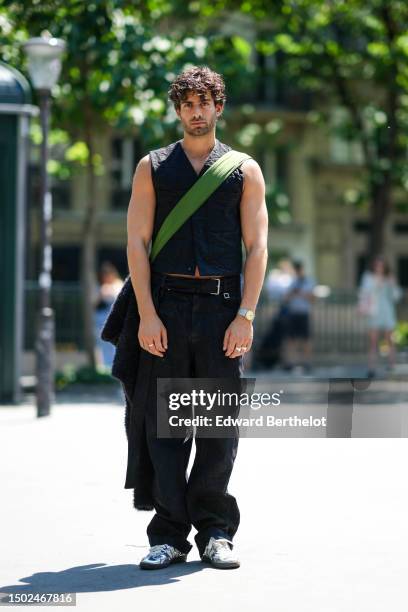 Guest wears a black sleeveless / buttoned gilet, a black shiny leather belt, black large denim pants, a green crossbody bag, silver shiny leather...