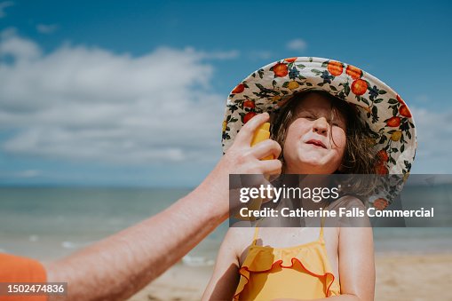 A parent applies sunscreen to a child's face - she braces as she prepares to be spritzed in the face A parent applies sunscreen to a child's face - she braces as she prepares to be spritzed in the face