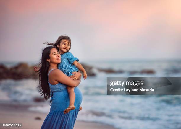 pregnant mother holding young daughter at the beach - mother and daughter in the wind stock-fotos und bilder